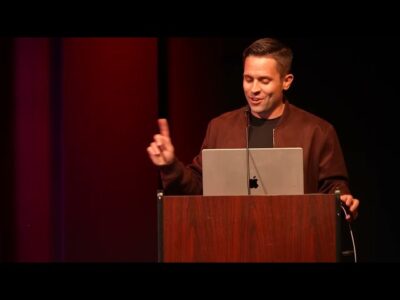 A man delivering a speech at a podium with a laptop displaying visual aids.