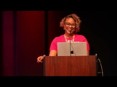 A woman in a pink shirt confidently stands at a podium, ready to address the audience.