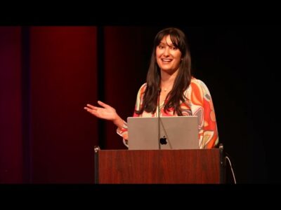 A woman delivering a speech at a podium with a laptop displaying visual aids.